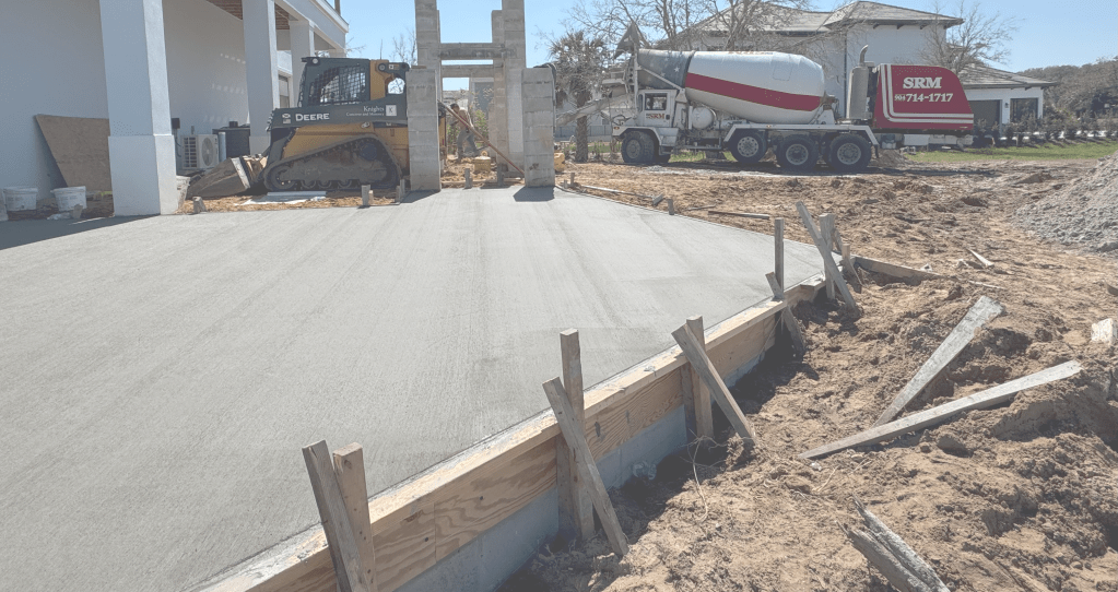 A construction site featuring a freshly poured concrete slab, with a bulldozer nearby and a concrete mixer truck in the background.