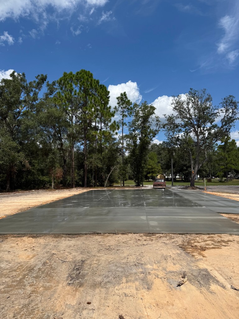 A newly poured concrete slab situated in a wooded area, with trees surrounding the site and a clear blue sky overhead.